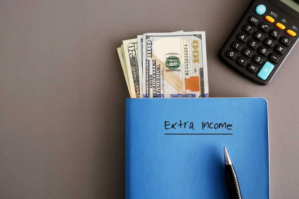 shutterstock 1552298207 Cash dollar money ,a pen, calculator on a blue notebook with text written on cover EXTRA INCOME, on grey background, concept of making more money with writing or freelance job gig worker